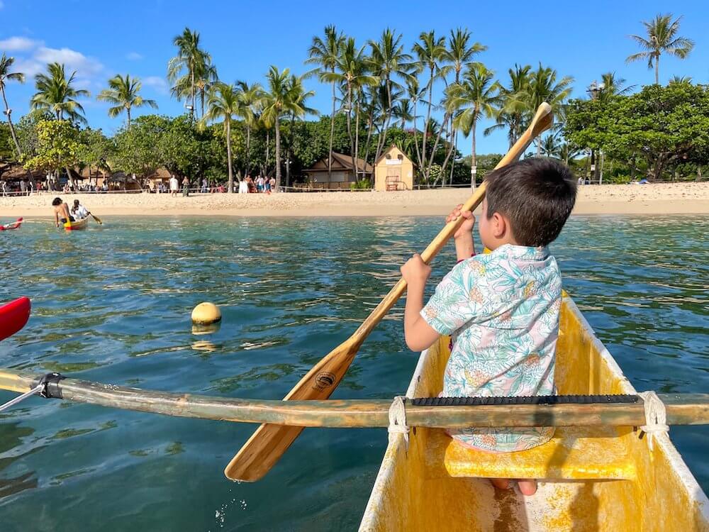 Image of a boy wearing an Aloha shirt paddling an outrigger canoe in a lagoon in Hawaii.