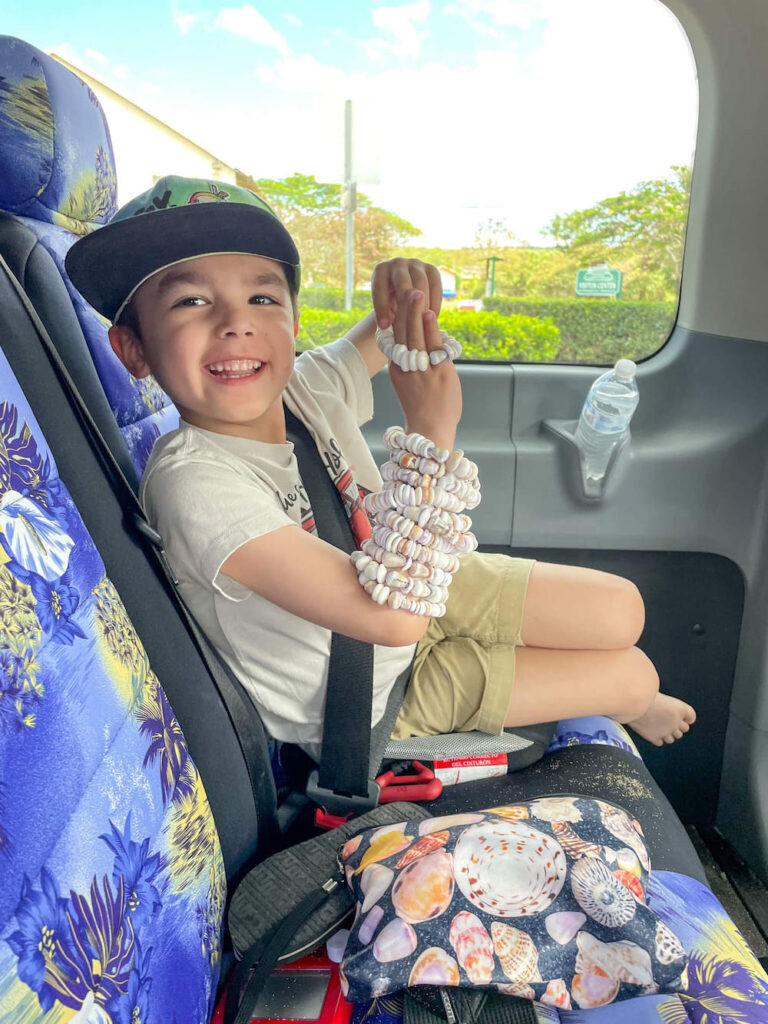 Image of a boy with a bunch of shell bracelets sitting on a booster seat inside a van on a circle the island tour in Oahu