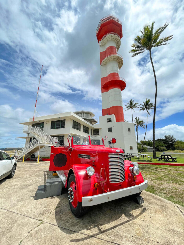 Image of a boy sitting in a vintage fire truck in front of a red and white striped control tower at Pearl Harbor