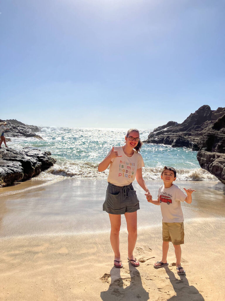 Image of a mom and boy waving shakas while standing on the beach of a small cove on Oahu