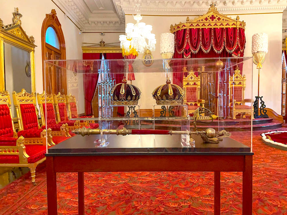 Image of a glass case with crowns in it and royal thrones in the background.