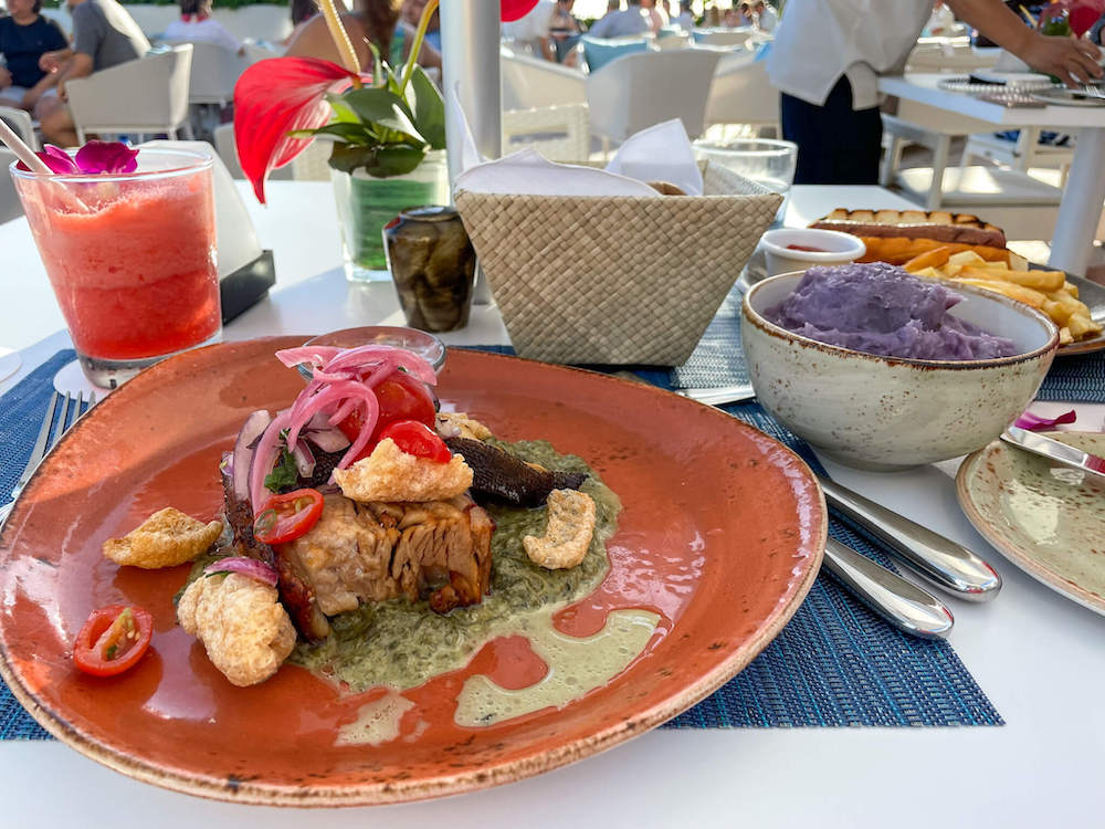 Image of a brown plate with upscale lau lau on it with a tropical pink cocktail and purple mashed potatoes