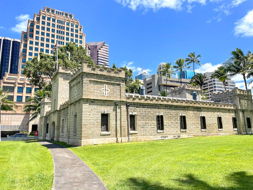 Image of a stone barracks on a grassy lawn surrounded by palm trees and buildings.