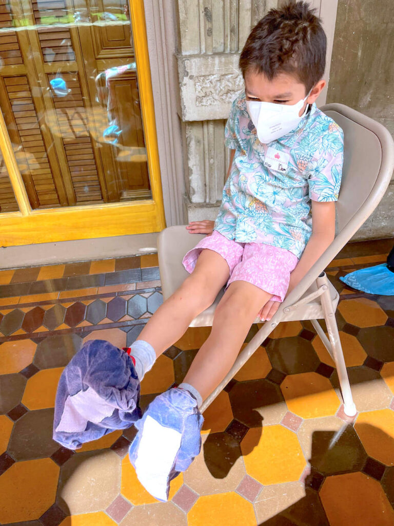 Image of a boy wearing an aloha shirt sitting in a chair with blue booties over his shoes.