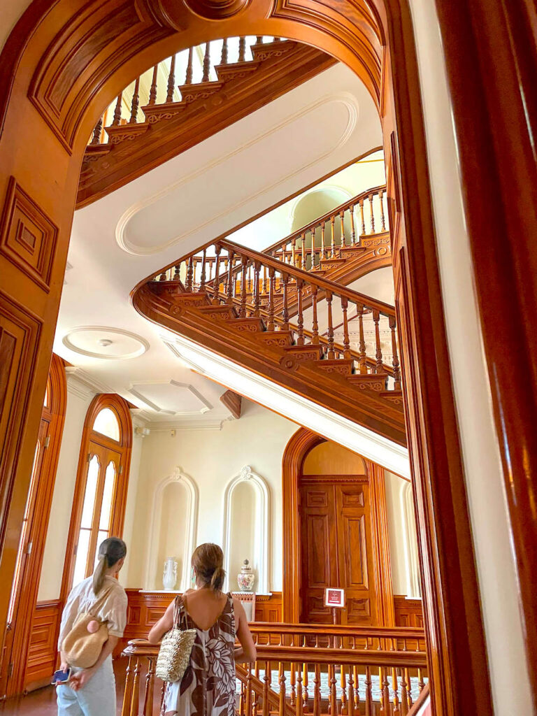 Image of two women standing along the railing of a gorgeous wooden staircase at Iolani Palace.