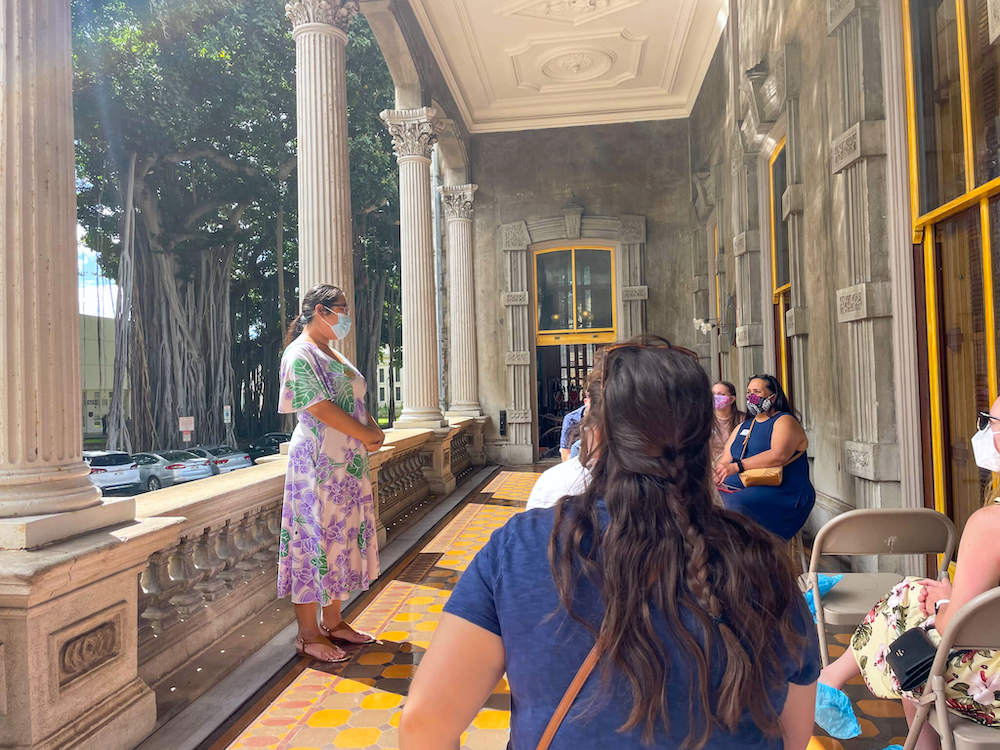 Image of a woman wearing a Hawaiian dress talking to a group of people sitting in chairs outside of Iolani Palace.