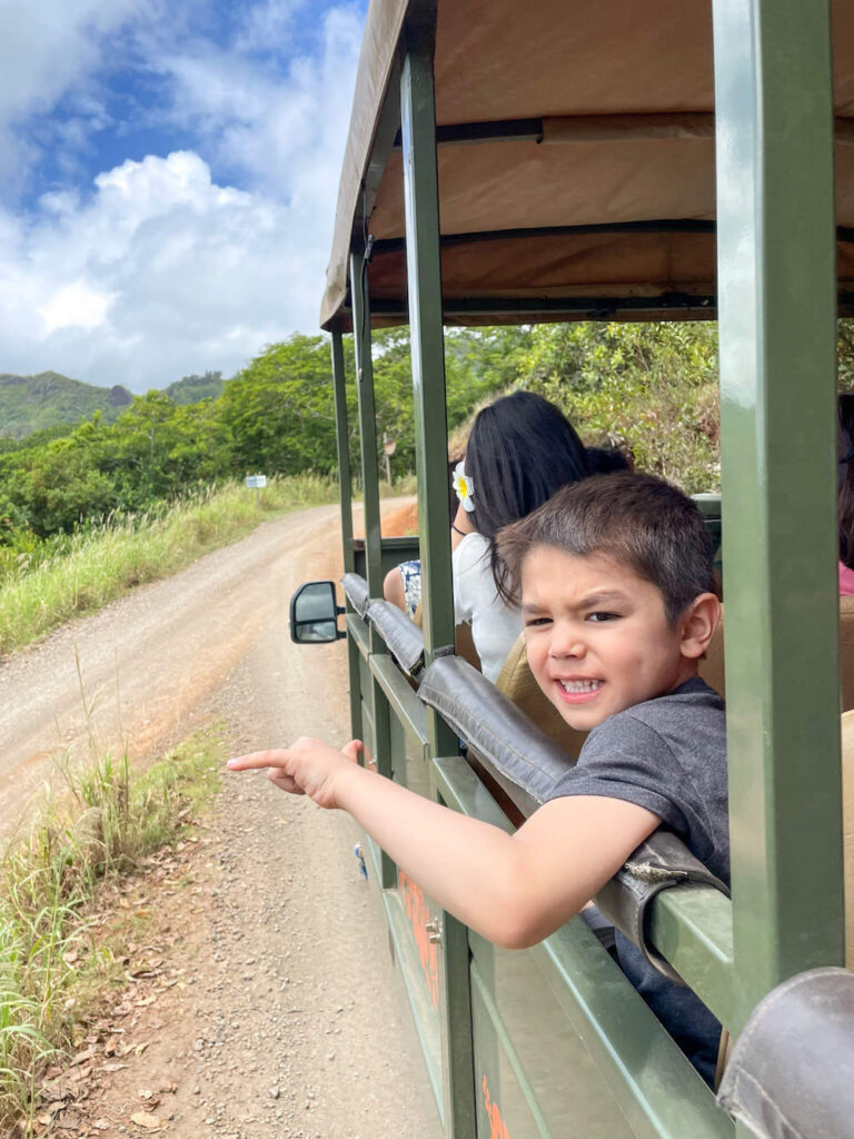 Image of a boy waving a shaka hand sign out the window of a Jeep at a ranch in Hawaii.