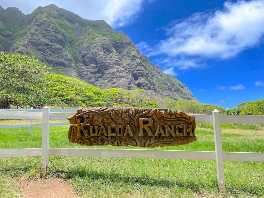 Image of a wooden Kualoa Ranch sign on a white fence with the Kualoa Mountains in the background