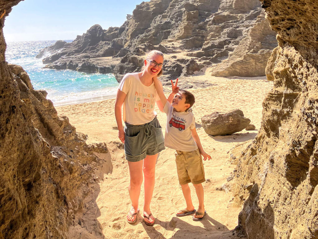 Check out this honest Oahu Circle Island Tour review by top Hawaii blog Hawaii Travel with Kids! Image of a mom and son at the opening of a lava tube with the ocean and beach in the background