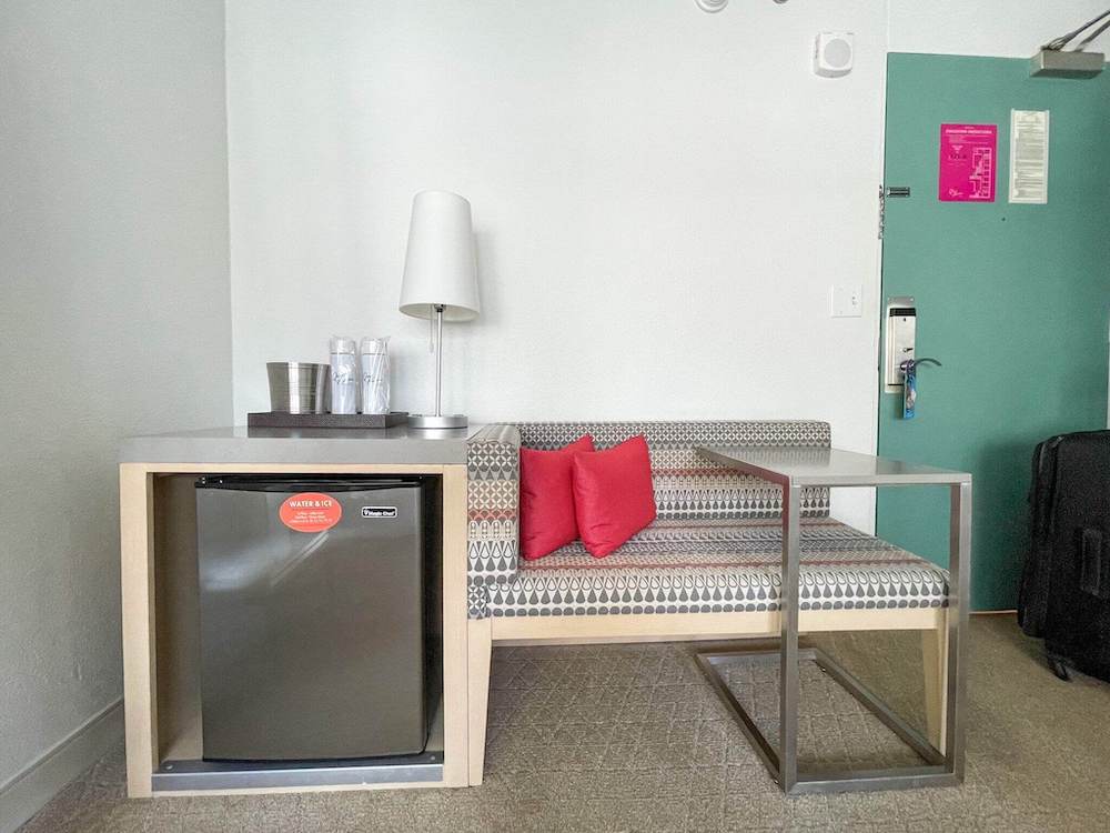 Image of a minifridge, bench seat, TV tray table, and seafoam green door inside the Queen Kapiolani hotel in Waikiki.