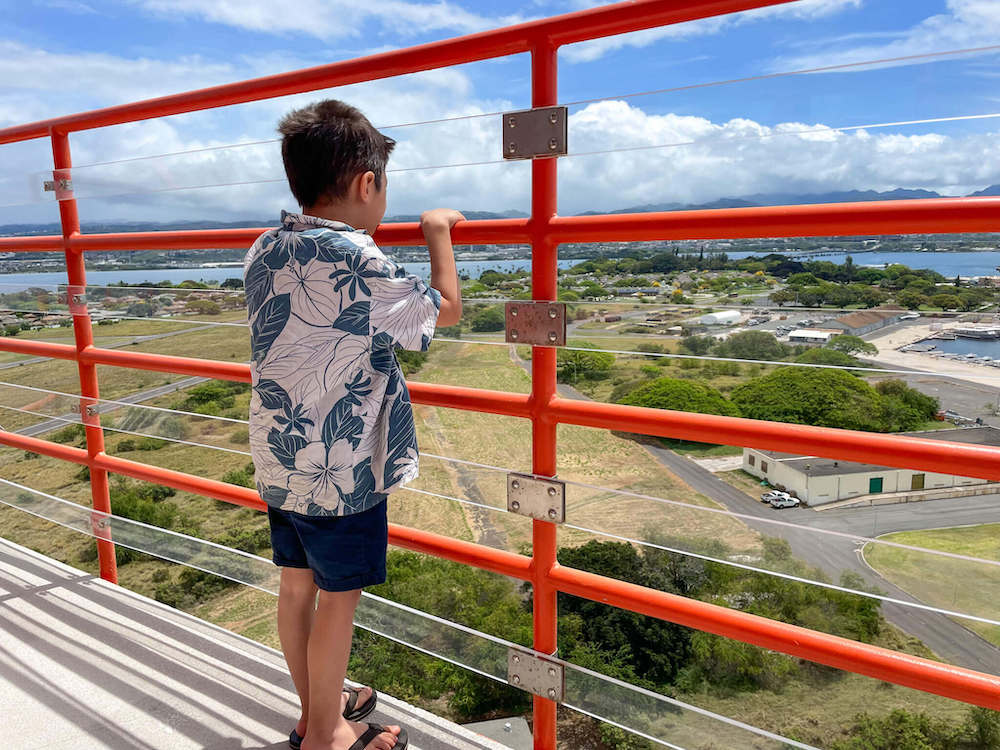 Image of a boy wearing an aloha shirt looking out between an orange railing out at Ford Island on Oahu