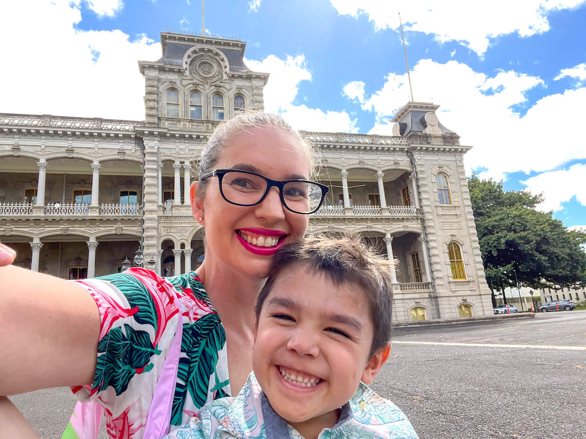Check out this guide to visiting Iolani Palace on Oahu by top Hawaii blog Hawaii Travel with Kids. Image of a mom and boy taking a selfie in front of an ornate building in Hawaii