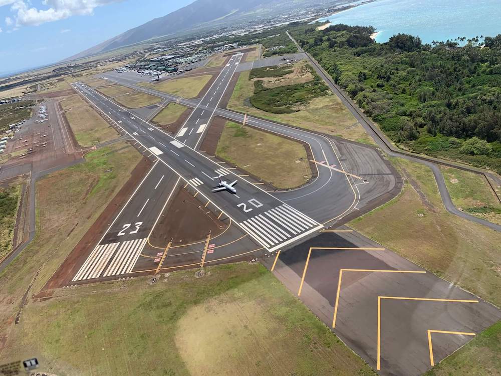 Image of an airport runway at Kahului Airport with green trees and the ocean surrounding it.