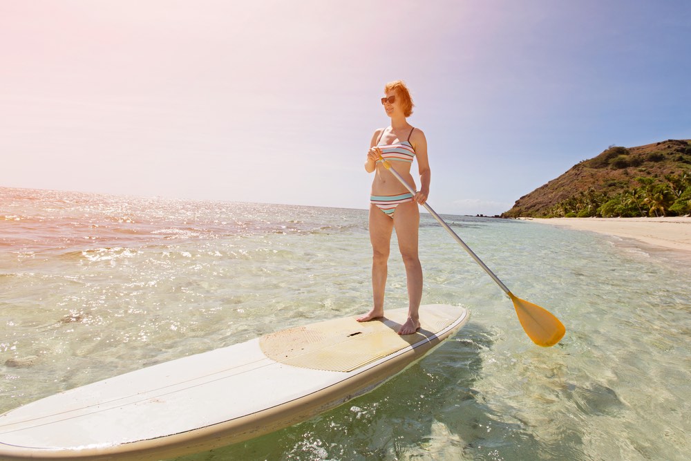 wide angle view of young woman enjoying stand up paddleboarding in beautiful turquoise lagoon at tropical island, active vacation concept, sun glare