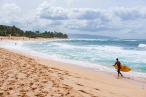 Find out the best things to do on Oahu in November recommended by top Hawaii blog Hawaii Travel with Kids. Image of Hawaii USA - November 7 2014; Young man carrying yellow surfboard along beach to go surfing.