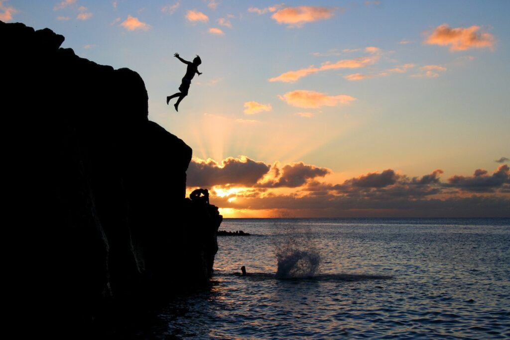 Image of a Boy jumps off a cliff into the ocean at Waimea Bay in Hawaii at sunset.