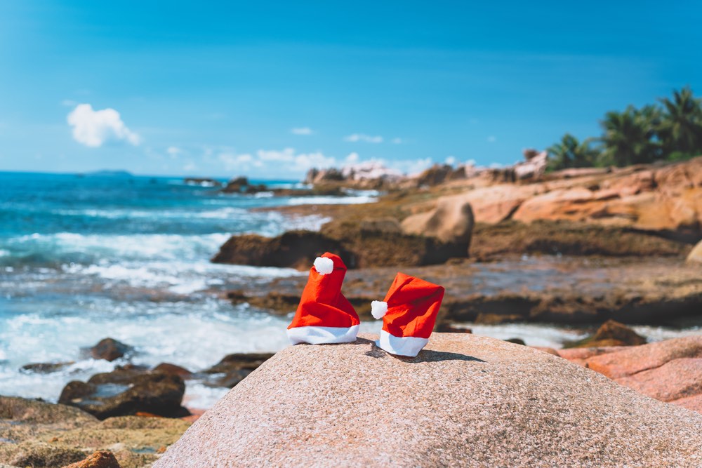 Couple of santa claus hat on tropical exotic paradise sandy beach with ocean waves and rocky coastline in background.