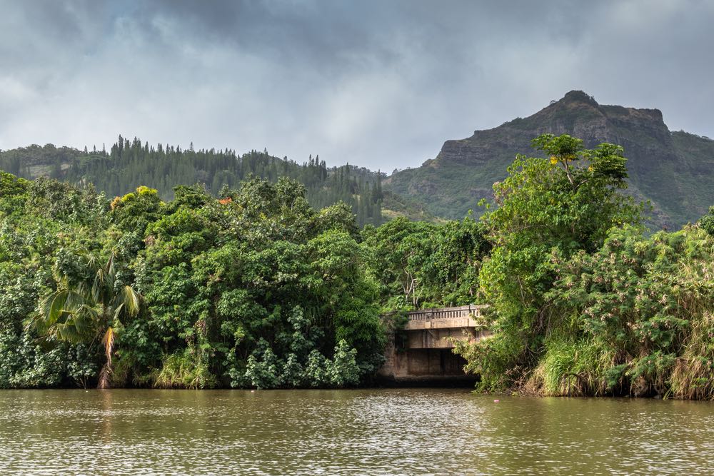 Nawiliwili, Kauai, Hawaii, USA. - January 16, 2020: Beige bridge over greenish South Fork Wailua River in front of green belt of trees under gray rainy cloudscape with brown rock mountain in back.