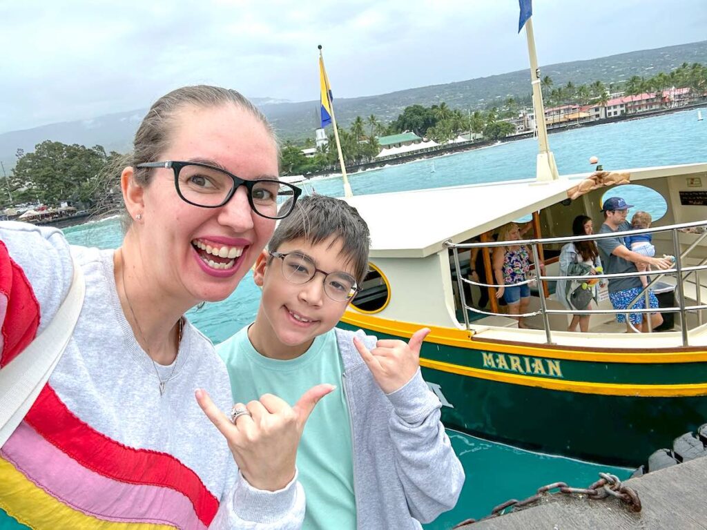Get the top tips and tricks for the Kona Glassbottom Boat Ride by top Hawaii blog Hawaii Travel with Kids. Image of a mom and son taking a selfie in front of the Kona Glass Bottom boat on the Big Island.