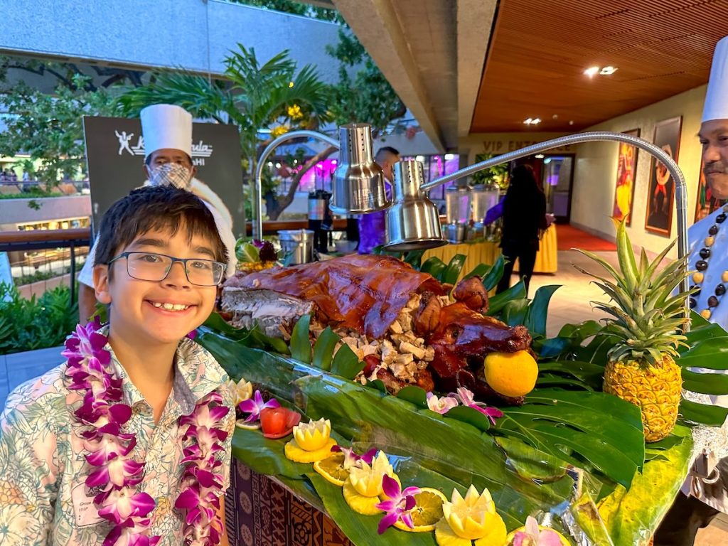 Image of a boy wearing a lei in front of a roasted pig