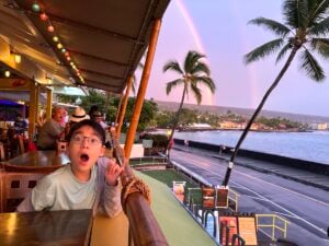 Image of a boy in a Kona restaurant with palm trees and a rainbow behind him