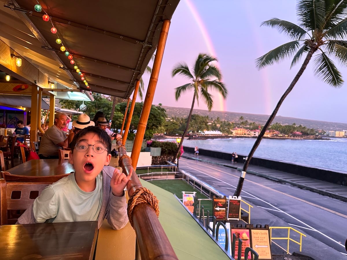 Image of a boy in a Kona restaurant with palm trees and a rainbow behind him